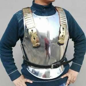May include: A polished steel breastplate with brass detailing and a black leather belt. The armour features decorative brass keyhole-shaped clasps and shoulder straps with a scalloped design. This historical piece is a functional and decorative item.