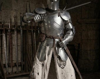 May include: A full suit of polished steel plate armor featuring a closed helmet with a cross-shaped visor, articulated gauntlets, and pauldrons. A white fabric tabard with a black eagle emblem is worn over the armor, secured by a brown leather belt.
