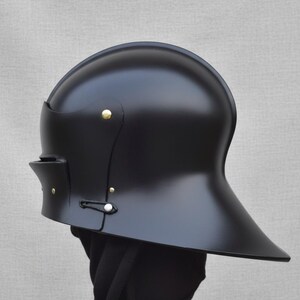 May include: A black, medieval-style helmet with a rounded top and a flared brim. The helmet features brass rivets and a chin strap. The surface has a glossy finish, and the helmet is displayed against a neutral background.