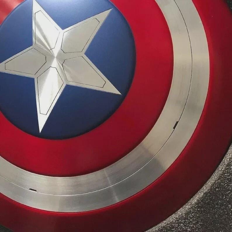 May include: A close-up of a Captain America shield. The shield is primarily red with a silver band and a blue circle in the center. A large, silver, five-pointed star is in the center of the blue circle. The shield has a metallic appearance.