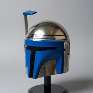 May include: A metallic silver and blue helmet with a black stand. The helmet has a blue visor and a silver dome. A blue antenna extends from the top. The helmet is a replica of a character's helmet from a science fiction film.
