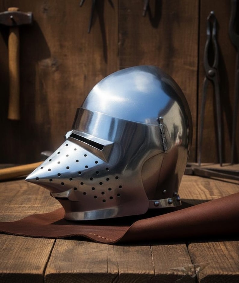 May include: A shiny, silver-coloured medieval helmet with a pointed visor and ventilation holes. The helmet rests on a brown leather surface, with a wooden background and blacksmith tools visible.