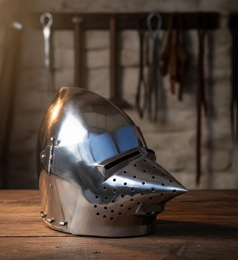 May include: A shiny, silver-coloured medieval helmet with a pointed visor and small ventilation holes. The metal helmet sits on a wooden surface. The helmet is a historical item, reflecting the light. The helmet is a historical item.