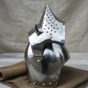 May include: A shiny steel medieval helmet, featuring a pointed visor with ventilation holes. The helmet has a rounded crown and a hinged neck guard. The polished metal reflects light, emphasizing the craftsmanship. The helmet is a historical item.