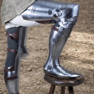 May include: Polished steel leg armor featuring articulated thigh, knee, and shin plates secured with brown leather straps and metal buckles. The knee section includes decorative spiral motifs, while the lower leg armor covers the foot with overlapping metal segments.