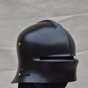 May include: A matte black helmet with a rounded top and a visor. The helmet has a small, curved brim and is adorned with two small, gold-colored rivets. The visor features a horizontal slit for vision. The helmet is likely for historical reenactment or costume use.