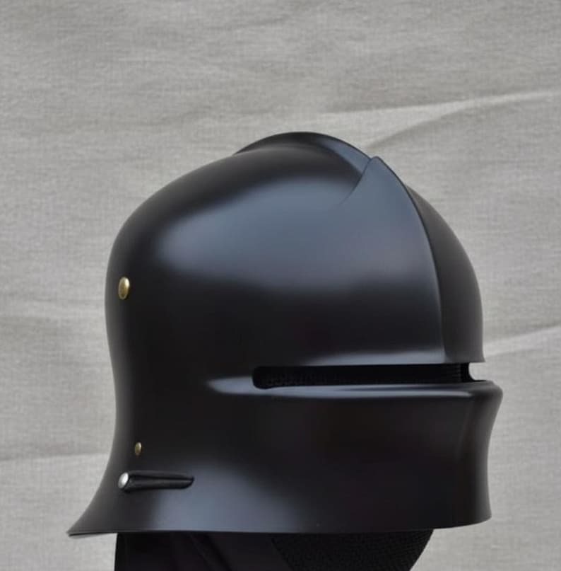 May include: A matte black helmet with a rounded top and a visor. The helmet has a small, curved brim and is adorned with two small, gold-colored rivets. The visor features a horizontal slit for vision. The helmet is likely for historical reenactment or costume use.