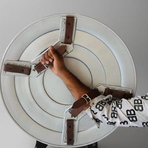 May include: A silver and white shield with brown leather straps and metal supports. The shield has a circular design with concentric rings and is held by a person's arm. The shield is a prop or costume accessory.