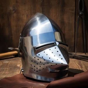 Puede incluir: Un yelmo medieval pulido, de color plateado, con visera puntiaguda y numerosos orificios de ventilación pequeños. El yelmo es de metal y tiene una superficie reflectante. Se encuentra sobre una superficie de cuero marrón, sugiriendo temas históricos o fantásticos.