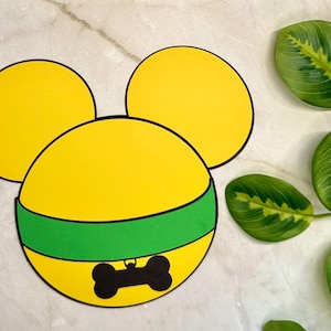 Puede incluir: Un diseño de estilo caricatura amarillo y verde con un contorno negro, que se asemeja a la cabeza de un personaje. El diseño incluye una banda verde y una forma de hueso negro. El fondo presenta hojas verdes con vetas amarillas.