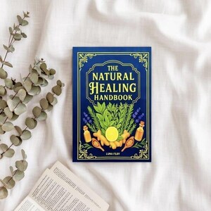 May include: A hardcover book titled "The Natural Healing Handbook" with a dark blue cover and gold lettering. The cover features illustrations of herbs, essential oil bottles, and a lemon. The book is on a white, textured surface.