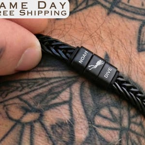 May include: A black braided leather bracelet featuring three rectangular metal beads. The center bead displays a white scuba diver icon, while the two flanking beads are engraved with the text NOAH and DIVE in a clean, sans-serif font.