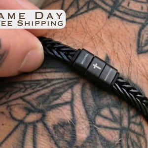 May include: A black braided leather bracelet featuring a rectangular metal clasp. The clasp is engraved with the name Michael, a small cross symbol, and the word Godfather. The bracelet has a textured, woven design and a smooth, dark finish.