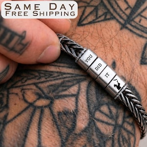 May include: Braided silver bracelet with three cylindrical beads. The beads read "YOU DID IT" and feature a small silhouette. The bracelet is on a tattooed arm.