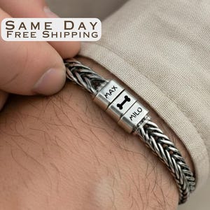 May include: Braided silver bracelet with a personalized clasp. The clasp has the names "MAX" and "MILO" engraved, along with a bone symbol. The bracelet is worn on a wrist.