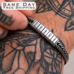 May include: A silver braided bracelet with personalized rectangular beads. The beads are engraved with the words "SINCE 1995", "JOHN", an infinity symbol, and "EMILY". The bracelet is on a wrist with tattoos.