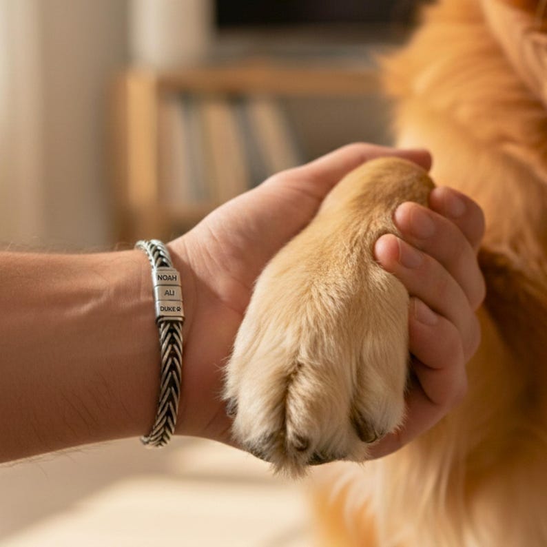 May include: A silver braided bracelet with the names "NOAH", "ALI", and "DUKE" engraved on it. The bracelet is on a person's wrist, holding a dog's paw. The dog has light brown fur.