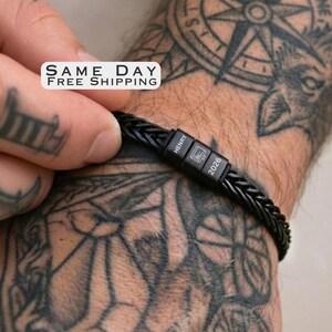 May include: A black braided bracelet featuring a rectangular metal clasp. The clasp is engraved with the name HENRY, a small computer monitor icon, and the year 2026. The bracelet has a textured, woven design and a matte black finish.