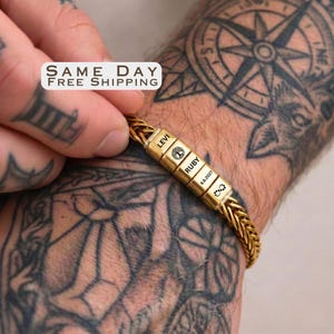 May include: A gold-toned wheat chain bracelet featuring four rectangular beads. The beads are engraved with the names LEVI and RUBY, a tree icon, a date 6.6.2027, and an infinity symbol. The bracelet is worn on a wrist with visible tattoos.
