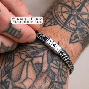 May include: A silver-toned braided chain bracelet featuring a rectangular metal clasp engraved with the name ERIC, a small silhouette of a person jumping, and the date 9.11.2026. The bracelet has a dark, oxidized finish within the chain links.