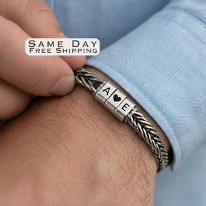 May include: A silver-toned braided chain bracelet featuring three cylindrical beads. The beads are engraved with the letter A, a heart symbol, and the letter E. The bracelet is worn on a wrist against a light blue fabric sleeve.