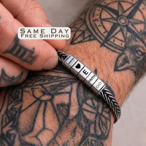 May include: A silver-toned metal bracelet featuring a braided chain design and four cylindrical beads. The beads are engraved with the text MMXXV, a heart symbol, the letter E, the word FAITH, and an infinity symbol in a dark, clear font.