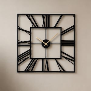 Op de afbeelding: Een vierkante zwarte metalen wandklok met Romeinse cijfers. De klok heeft een vierkant midden met een zwarte wijzerplaat en goudkleurige wijzers. De klok is bevestigd aan een lichtgekleurde muur in een woonkamer.