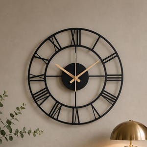 May include: A large circular wall clock with a black metal frame and Roman numerals. The clock features a central black circular plate with thin gold-coloured hour and minute hands. The design is open-faced, showcasing the wall behind the clock.