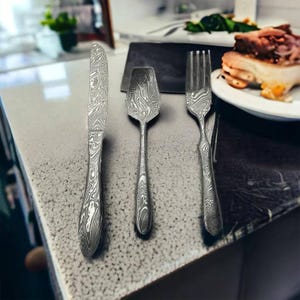 May include: A set of three stainless steel utensils: a knife, spoon, and fork. The utensils have a unique patterned design. The knife is on the left, the spoon in the middle, and the fork on the right. The utensils are on a speckled countertop.