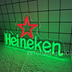 May include: A neon sign with the word "Heineken" in bright green, with a red star above the text. The sign is on a clear acrylic backing. The text "KETCHANATSHOP" is below the word "Heineken".