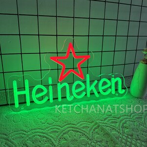 May include: Green neon sign with the word "Heineken" and a red star. The sign is on a clear backing and has the text "KETCHANATSHOP" below the word "Heineken". The background is a green grid.