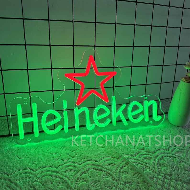 May include: Green neon sign with the word "Heineken" and a red star. The sign is on a clear backing and has the text "KETCHANATSHOP" below the word "Heineken". The background is a green grid.