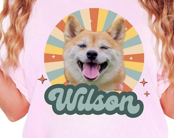 May include: A light pink t-shirt featuring a smiling Shiba Inu dog with a retro-style design. The dog is set against a circular background with orange, yellow, and teal rays. The name "Wilson" is written in a green, cursive font below the dog.