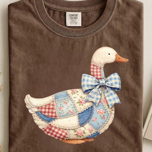 Puede incluir: Una camiseta marrón Comfort Colors con un diseño de ganso de patchwork. El ganso tiene una cabeza blanca, pico naranja y un lazo de cuadros azul. El patchwork incluye patrones florales y de cuadros. La camiseta tiene cuello redondo y una etiqueta "Comfort Colors".