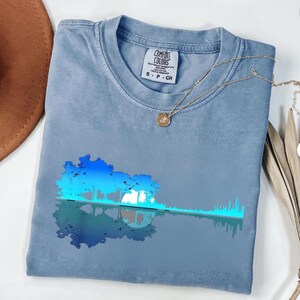 以下が含まれることがあります： ギターの形をしたグラフィックが特徴の青いComfort Colors Tシャツ。木と水の反射のデザインです。シャツはクルーネックと半袖です。ペンダント付きのゴールドネックレスがシャツの上にかけられています。