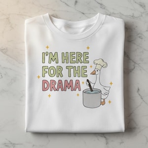 Puede incluir: Camiseta blanca con el texto "I'M HERE FOR THE DRAMA" en verde y rosa. Un dibujo de un ganso con gorro de chef removiendo una olla. Estrellas amarillas adornan el texto y el ganso.