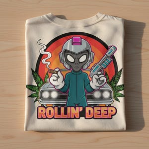 May include: Cream-colored t-shirt featuring a cartoon alien with a helmet, holding a cigar and a futuristic gun. The alien is in front of a car with a rainbow background and the text "ROLLIN' DEEP".