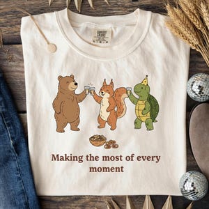 Puede incluir: Camiseta color crema con un oso, una ardilla y una tortuga de dibujos animados brindando con tazas. Debajo hay un cuenco con pretzels. El texto "Making the most of every moment" está impreso debajo.