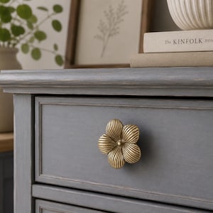May include: A gray wooden dresser drawer featuring a brass-toned metal knob shaped like a five-petaled flower with ribbed texture. The drawer front has a rectangular recessed panel design and a smooth, matte finish on the painted wood surface.