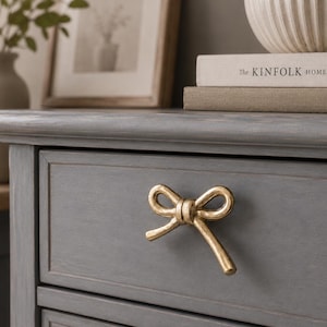 May include: A gold-toned metal drawer pull shaped like a tied bow, mounted on a gray wooden drawer front. The bow features a textured, metallic finish with two loops and two trailing ends, providing a functional handle for the furniture piece.