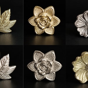 May include: A collection of six metal pins featuring botanical and floral designs. The set includes two leaf-shaped pins, two rose-shaped pins, and two daisy-shaped pins, available in both gold-toned and silver-toned metallic finishes.