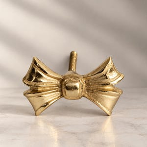 Peut inclure: Un bouton de meuble en métal doré en forme de nœud. Le nœud présente un nœud central carré avec deux boucles plissées et évasées s'étendant sur les côtés. Une tige filetée en métal dépasse du centre arrière de cette pièce de quincaillerie.