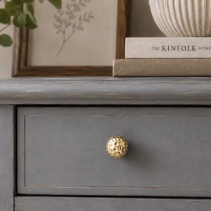 Peut inclure: Un bouton de meuble rond de couleur dorée avec une surface texturée et martelée. Le bouton est fixé sur une façade de tiroir en bois gris présentant un design à panneau encastré rectangulaire avec un grain de bois visible et des bords usés.