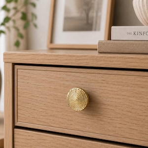May include: A round, gold-colored metal cabinet knob featuring a textured, concentric spiral pattern. The knob is mounted on the front of a light-toned wooden drawer, which displays a visible wood grain and a simple, clean-lined rectangular frame.