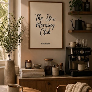 Puede incluir: Un cuadro de pared enmarcado que presenta el texto negro The Slow Morning Club en letra cursiva, con el mismo texto repetido en una fuente sans-serif más pequeña debajo. La impresión se muestra en un marco de madera sobre una pared.