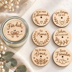 Könnte beinhalten: Eine Glaskerze mit einem runden Holzdeckel, der mit dem Text Für dich und einer Schleife graviert ist, sowie sechs weiteren runden Holzscheiben mit verschiedenen deutschen Sprüchen wie Lieblingmensch, Mama, Oma und Beste Freundin.