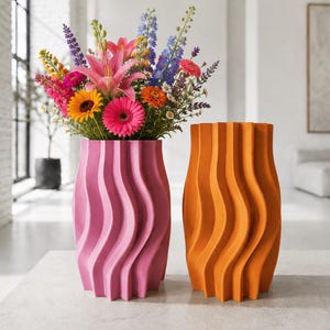 Könnte beinhalten: Zwei zylindrische Vasen mit einer vertikalen, wellenförmigen Rippenstruktur. Eine Vase ist mattrosa und enthält einen bunten Strauß aus rosa, orangefarbenen, gelben, violetten und blauen Blumen. Die zweite Vase ist mattorange und steht leer daneben.