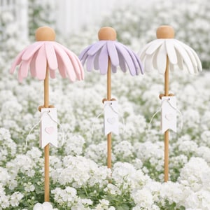 Op de afbeelding: Drie decoratieve bloementuinpalen met roze, paarse en witte bloemblaadjes. Elke paal heeft een houten steel, een kleine houten bal aan de bovenkant en een wit label met een hart. De palen staan in een bed van witte bloemen.