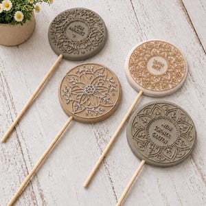 May include: Four circular decorative plaques on wooden sticks, featuring intricate embossed floral and mandala patterns. Two plaques are grey, one is beige, and one is white. The grey plaques feature the German text "MEIN PARADIES" and "MEIN SCHÖNER GARTEN".