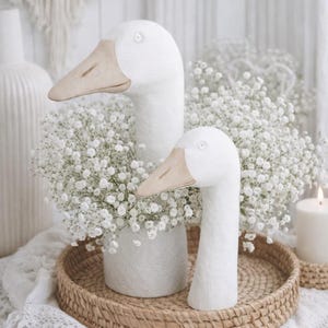 Könnte beinhalten: Zwei weiße Gänse-Skulpturen mit beigen Schnäbeln, eine hohe und eine kürzere, stehen auf einem gewebten Tablett. Die Gänse sind von weißen Schleierkrautblüten in einer weißen Vase umgeben. Im Hintergrund brennt eine Kerze.